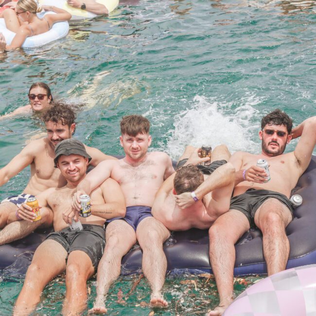 Six young men in swimwear relax on a large inflatable mattress in a pool, holding drinks and smiling. Other people on floaties are in the background, enjoying the sunny, festive atmosphere.