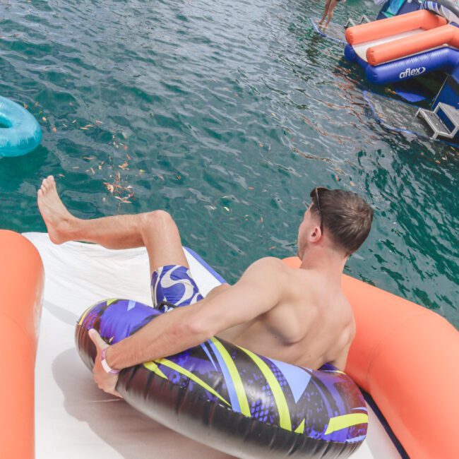 A man in swim trunks sits on an inflatable tube at the top of a slide, ready to go into a lake with boats and other people on floats in the background.