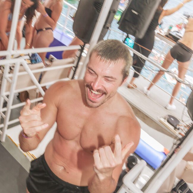 A smiling man in swim trunks makes rock-and-roll hand signs on a boat deck, surrounded by people in swimwear enjoying a party. The atmosphere is lively and energetic, with the sea visible in the background.