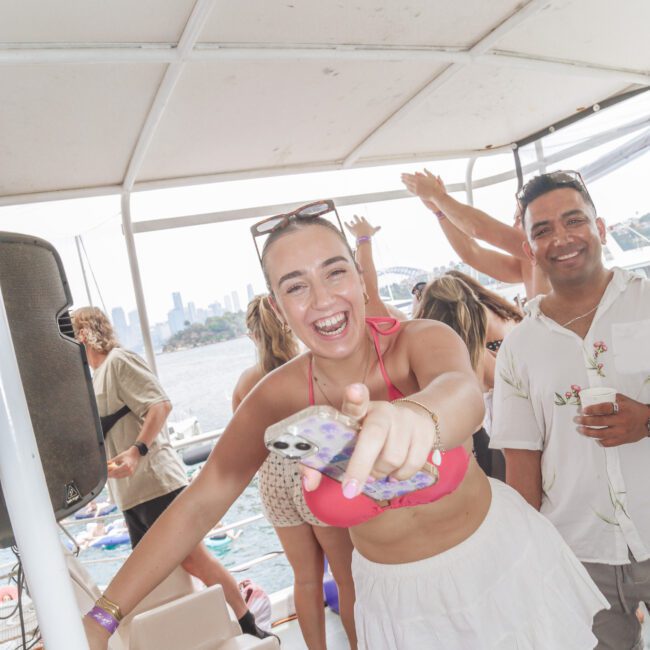 A woman in a pink bikini top and white skirt smiles and points towards the camera while holding a phone at a lively boat party. Other people are dancing and mingling in the background on a sunny day by the water.