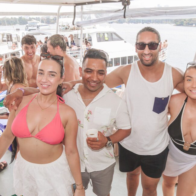 Four people smile and pose together on a boat, with others and additional boats visible in the background on the water, creating a lively, sunny party atmosphere.