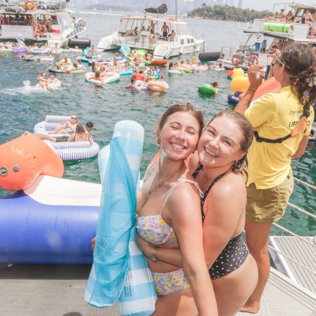 Two young women in swimsuits smile and pose on a boat, holding a pool float. Behind them, people enjoy a lively party with inflatables in the water and on several boats under a sunny sky.
