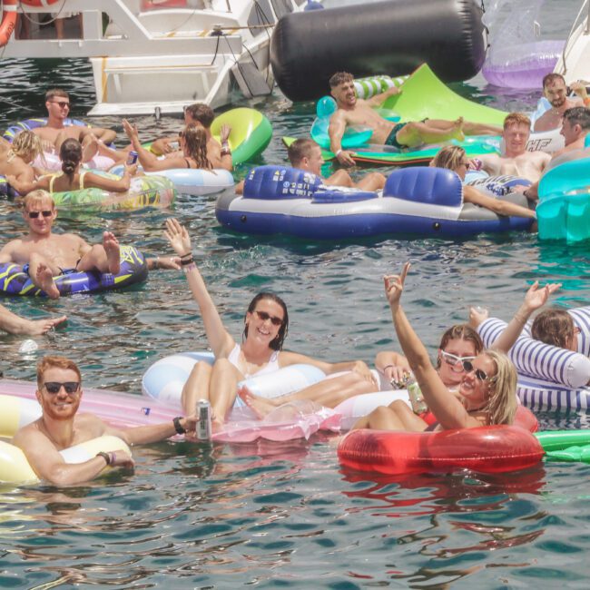 A group of people relax on colorful inflatable floats in the water, smiling and waving at the camera during a sunny day. Boats are docked nearby, and the atmosphere is lively and festive.