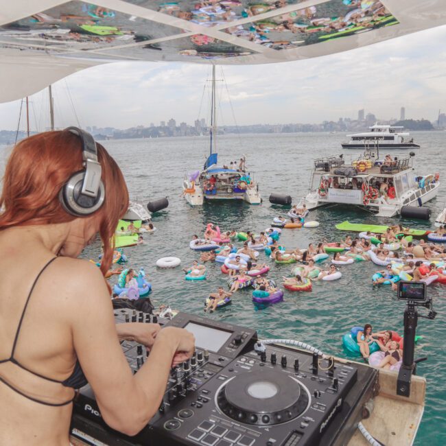 A female DJ wearing headphones plays music on a boat, overlooking a lively crowd of people in swimwear floating on inflatables and boats in a bay, with city buildings visible in the background.