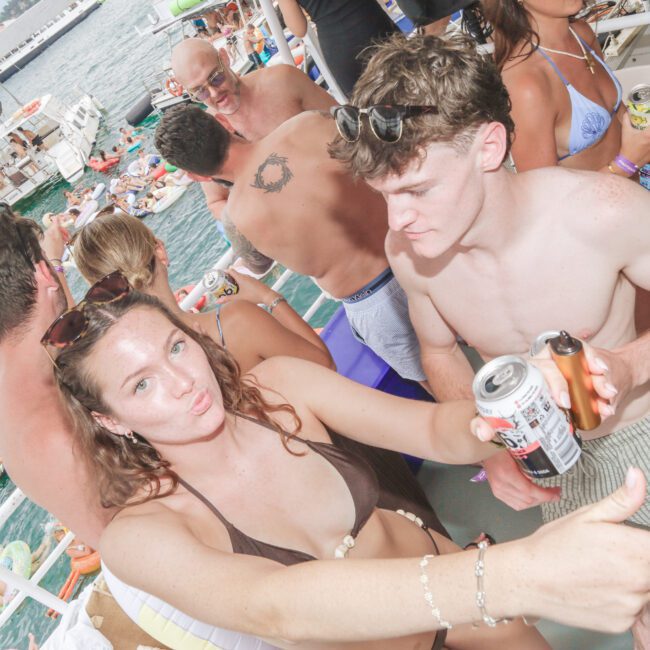 A group of young adults in swimsuits are partying on a boat, holding drinks and dancing. People in the background are floating on the water with inflatables. The atmosphere is lively and festive, under a partly cloudy sky.