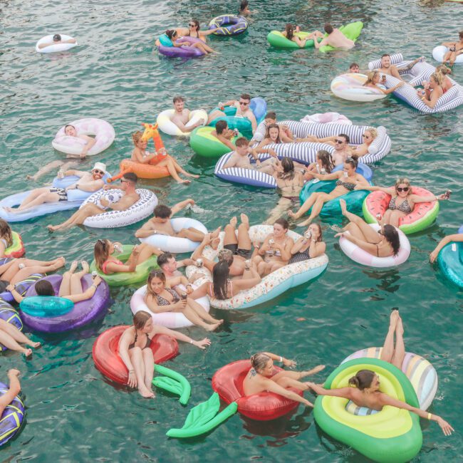 A large group of people relax on colorful inflatable rafts and pool floats in the water, enjoying a lively outdoor social event under sunny weather.