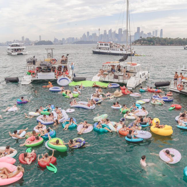 A large group of people relax on colorful inflatables in the water beside several boats, with a city skyline and bridge visible in the background on a partly cloudy day.