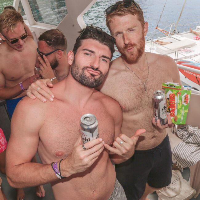 Two shirtless men pose on a boat, holding drinks and a beer box, smiling at the camera. Other people in swimsuits are in the background. The boat is docked near clear water and another orange boat is visible outside.