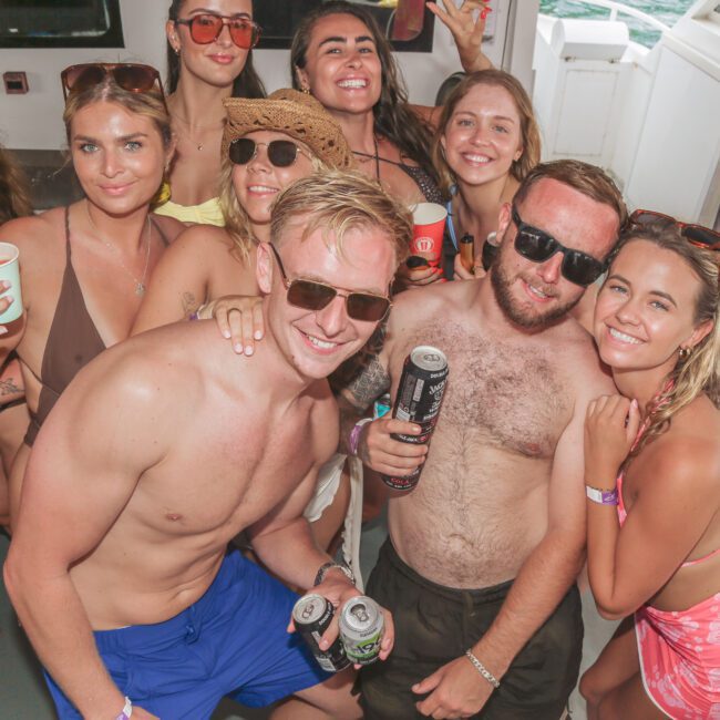 A group of young adults in swimsuits, some wearing hats and sunglasses, smile and pose together on a boat, holding drinks and enjoying a sunny day.