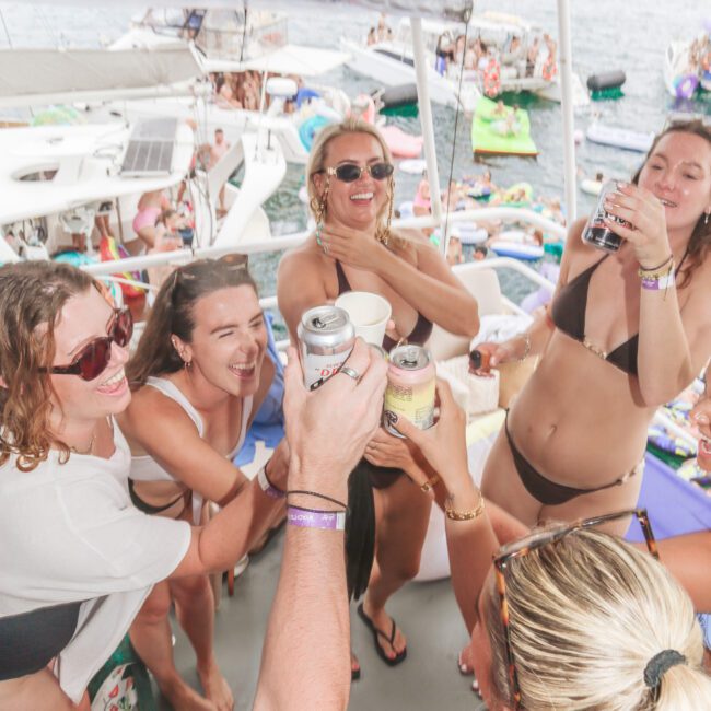 A group of women in swimsuits laughing and raising drinks together on a boat, surrounded by other boats and people on the water, with a festive, summery atmosphere.