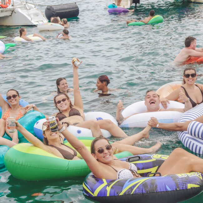 A group of people relax and smile on colorful inflatable tubes in the water, holding drinks, surrounded by others floating nearby. Boats and more people are visible in the background, enjoying a lively day on the water.
