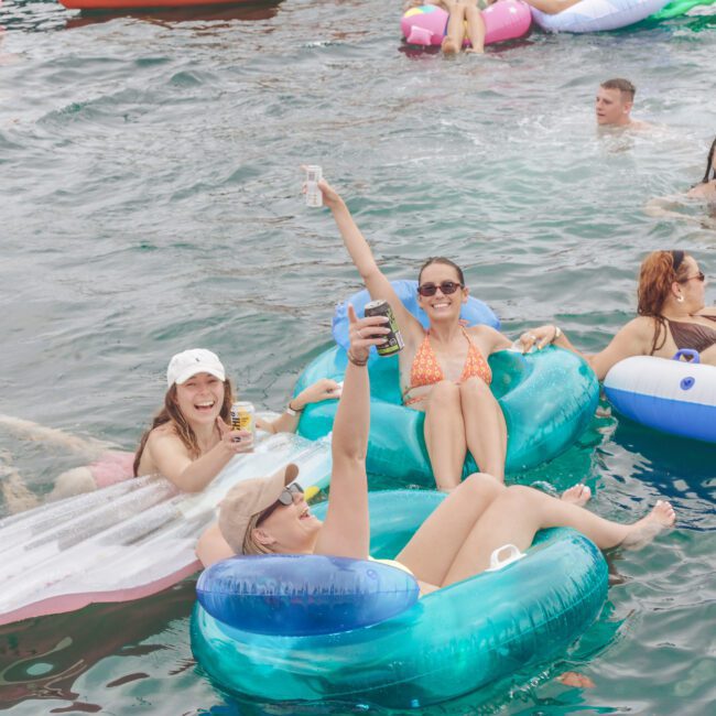 A group of people relax on colorful inflatable floats in a lake, smiling and holding drinks. The water is clear, and everyone appears to be enjoying a sunny day outdoors.