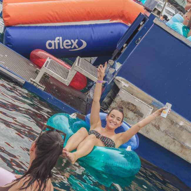 A woman in a polka dot bikini relaxes on a turquoise pool float, smiling and raising her arms in celebration, while another person floats nearby at a lively yacht party. Inflatable toys and water are visible around them.