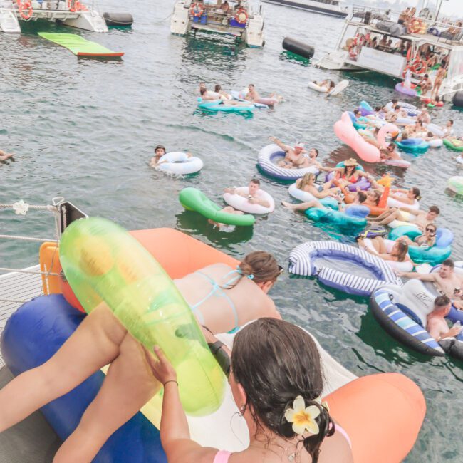 A crowded scene on the water with people relaxing on inflatable floats and boats. In the foreground, a woman with a flower in her hair helps another person onto a large inflatable from a yacht. The atmosphere is lively and festive.