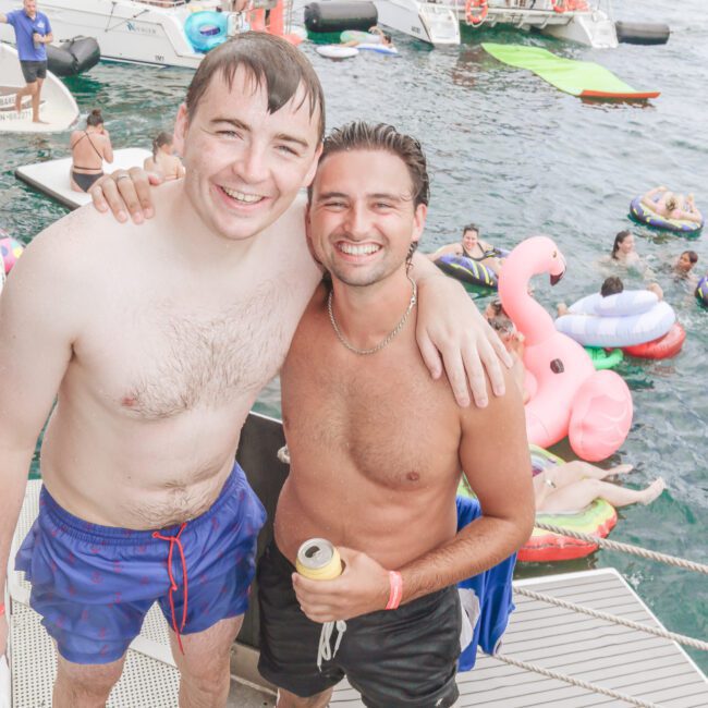 Two smiling men in swim trunks stand arm in arm on a boat ramp, with one holding a drink. Behind them, people relax on boats and colorful pool floats in the water during a lively party.
