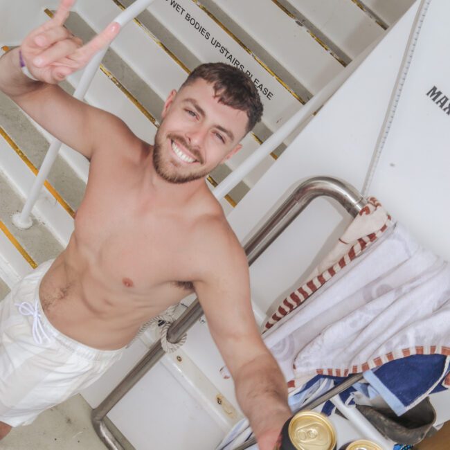 A smiling shirtless man in white shorts stands on a boat staircase, holding two cans of beer and flashing a peace sign. Towels hang on the rail beside him. "Yacht Social Club" logo is visible in the corner.