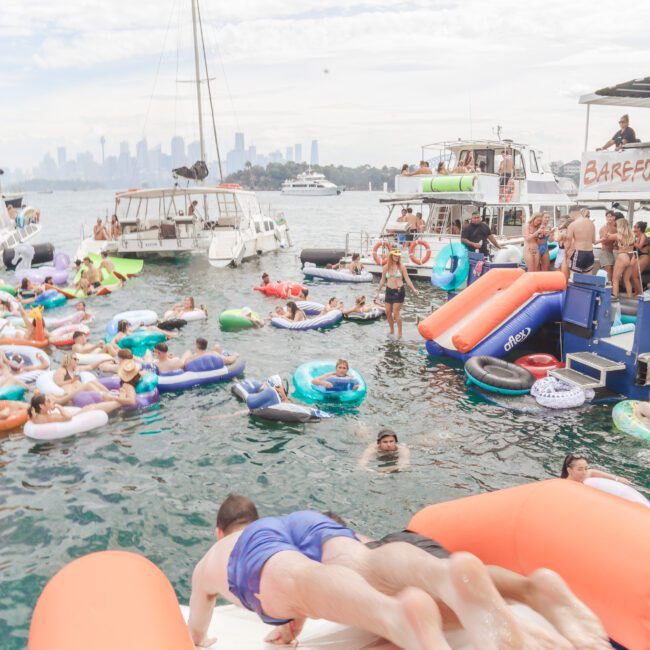 People relax on colorful pool floats in the water near several boats. In the foreground, two people slide down an inflatable water slide toward the water. The background shows a city skyline under a cloudy sky.