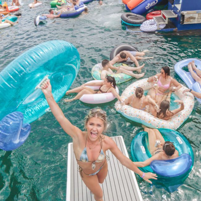 A smiling woman in a bikini stands on a dock, raising her arm, while people float on colorful inflatables and relax in the water around her on a sunny day.