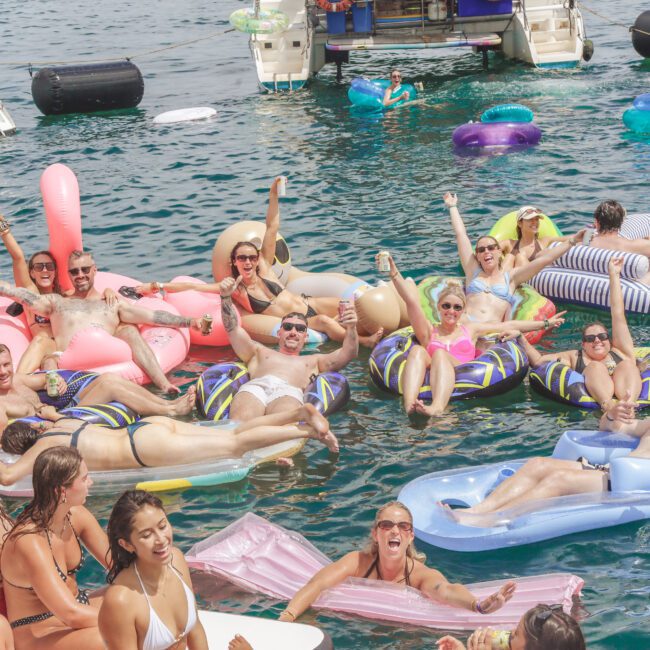 A group of people relax on colorful pool floats in the water near boats, smiling and enjoying a sunny day. Some hold drinks, and everyone appears cheerful and festive.