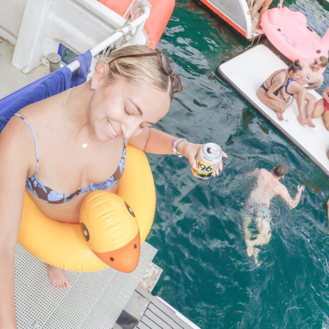 A smiling woman in a bikini and yellow duck float holds a drink on a boat, with people lounging and swimming in turquoise water below on a sunny day.