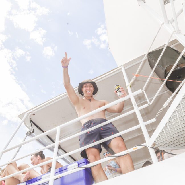 A smiling man in swim trunks and a hat stands on a boat deck, holding a can and pointing upward. Other people sit nearby, relaxing. The sky is bright and partly cloudy above.
