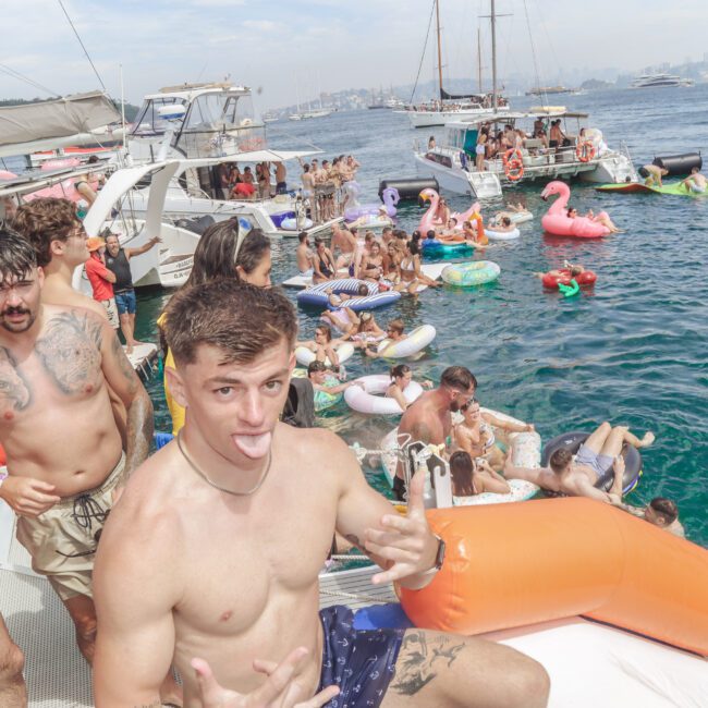 A group of young people on a boat and in the water at a lively pool party, with inflatables, yachts, and city skyline in the background. One man in front poses playfully, sticking out his tongue.