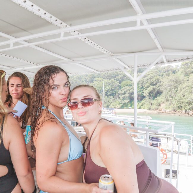 Several women in swimsuits are gathered on a boat, with two women in the front posing closely and making playful faces for the camera. The boat is on water, and lush green trees are visible in the background.