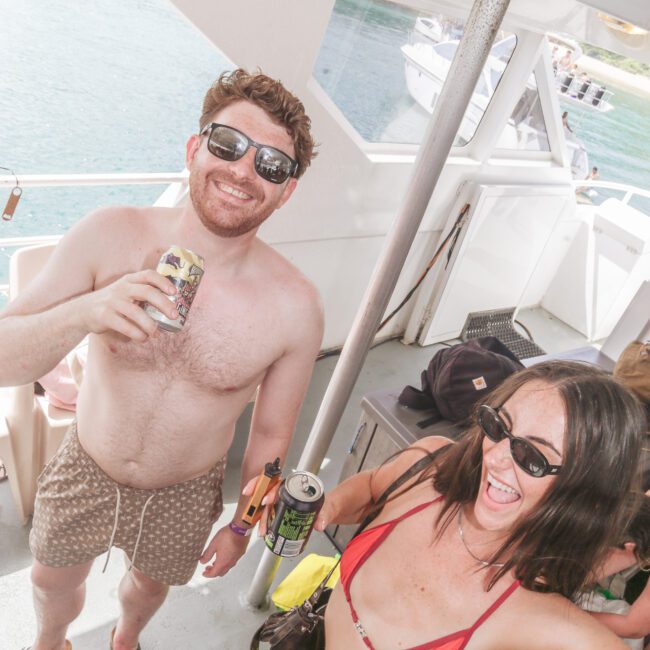 A man in swim trunks and sunglasses holds a drink and smiles next to a woman in a red bikini and sunglasses, also holding a drink, on a boat with water and other boats visible in the background.