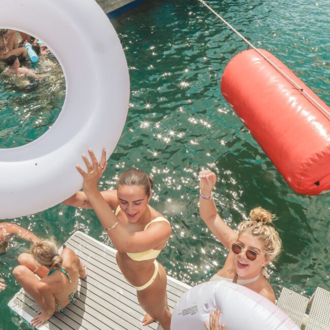 Two women in swimsuits stand on a dock by the water, smiling and holding large inflatable rings. Other people swim nearby. The sun is shining, and the mood is lively and fun.