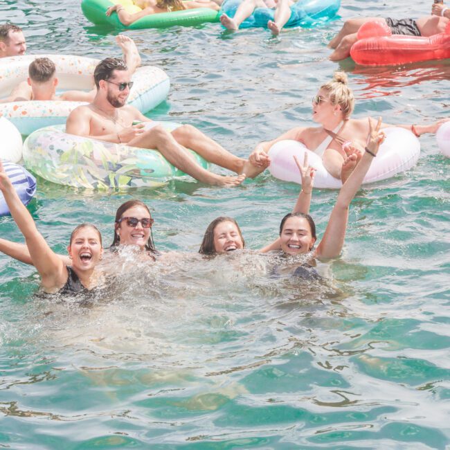 Four women in swimsuits smile and raise their arms while standing in clear water. Behind them, people relax on colorful inflatable floats, enjoying a sunny day on the water.