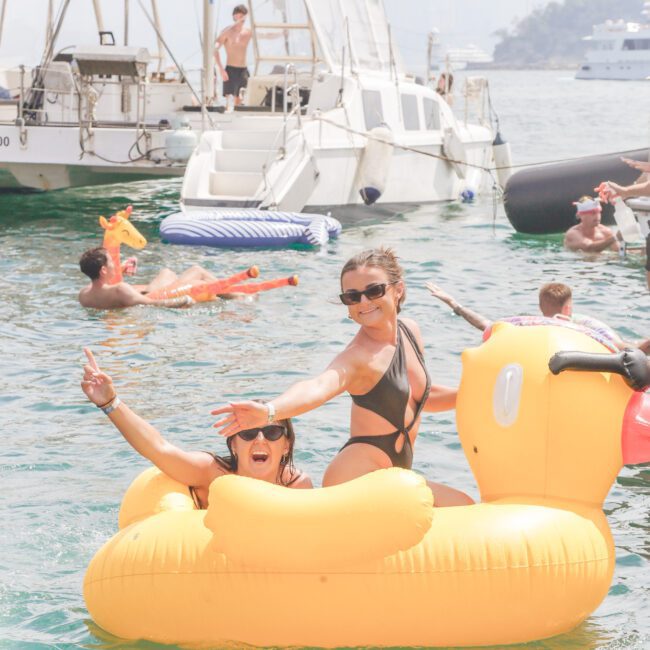 Two women in swimsuits smile and pose on a large yellow duck float in the water, surrounded by boats and other people enjoying a sunny day. City buildings appear in the hazy background.