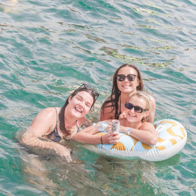 Three women in swimsuits smile at the camera while enjoying the water; one is on an inflatable pool float, and two are in the water. They all wear sunglasses. Other swimmers and floats are in the background.