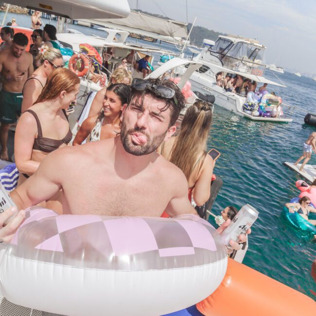 A man holding a pink-and-clear swim ring squints at the camera on a crowded boat party, surrounded by people in swimwear, with other boats and blue water in the background.