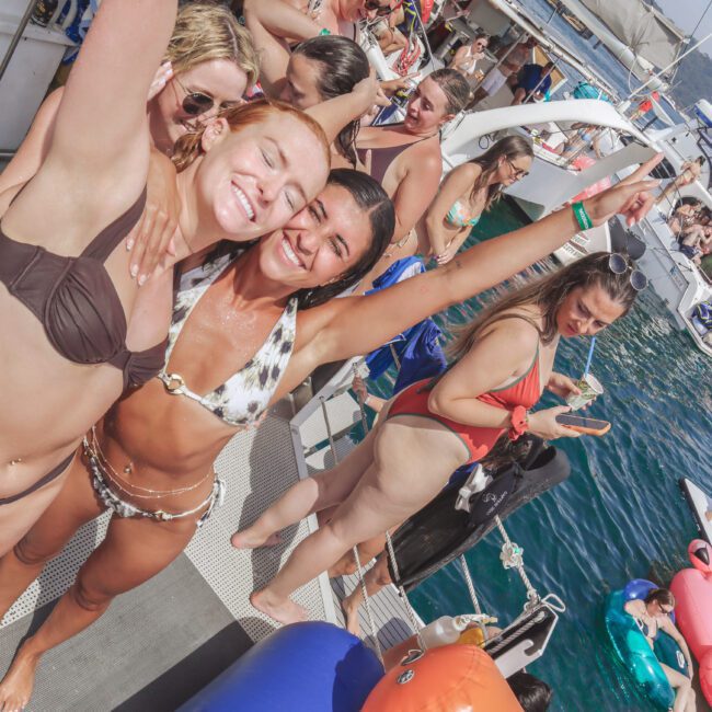 Two young women in swimsuits smile and pose with arms raised on a crowded boat, surrounded by other people enjoying a sunny day on the water. Inflatable floats and the blue sea are visible in the background.