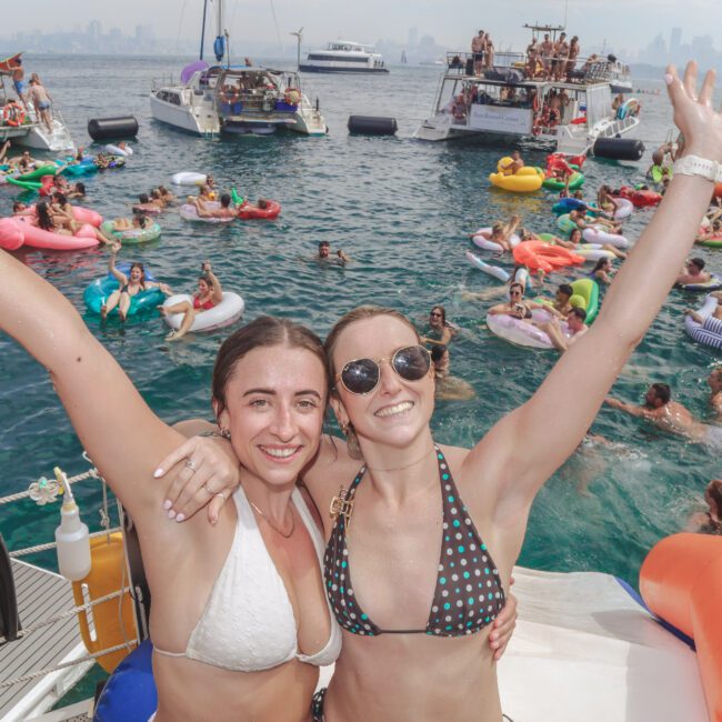 Two women in bikinis pose and smile with arms raised on a boat, surrounded by people swimming, floating on inflatables, and relaxing on other boats in the water, with a city skyline visible in the background.