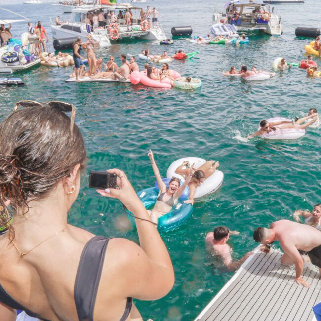 A woman on a boat takes a photo of people swimming and relaxing on inflatables in a crowded, lively water party scene with several boats in the background.