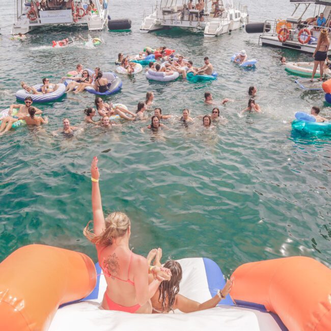 People enjoying a summer party in the water with inflatables, slide, and boats in the background. Two women sit on an inflatable slide at the front, with one waving to the crowd. The scene is lively and festive.