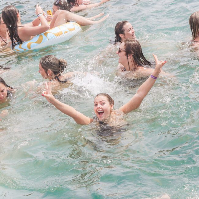 A group of people are swimming and having fun in clear blue water. One person in the center is smiling with arms raised, making peace signs with both hands. Others are laughing and talking nearby. An inflatable float is visible.