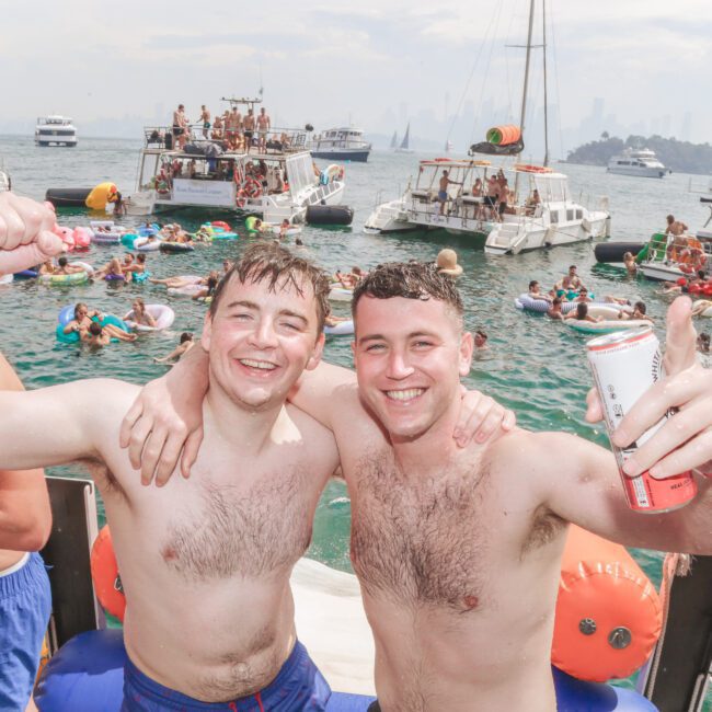 Two smiling young men stand shirtless on a boat, arms around each other, holding drinks. Behind them, people swim and lounge on inflatables in the water, with several boats and a city skyline in the background.