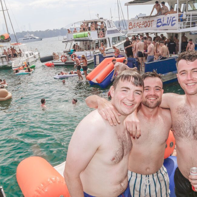 Three smiling young men in swim trunks pose together on a boat at a lively water party, with people swimming and relaxing on inflatables nearby and several boats anchored close together in the background.