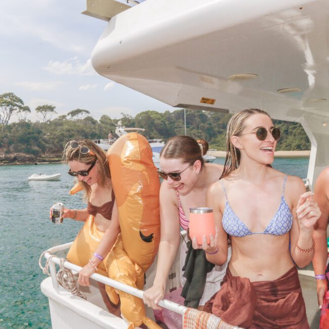 Four women in swimsuits laugh and enjoy drinks on a boat, one wearing an inflatable dinosaur costume. They are docked near a scenic, tree-lined shore with calm blue water and other boats in the background.