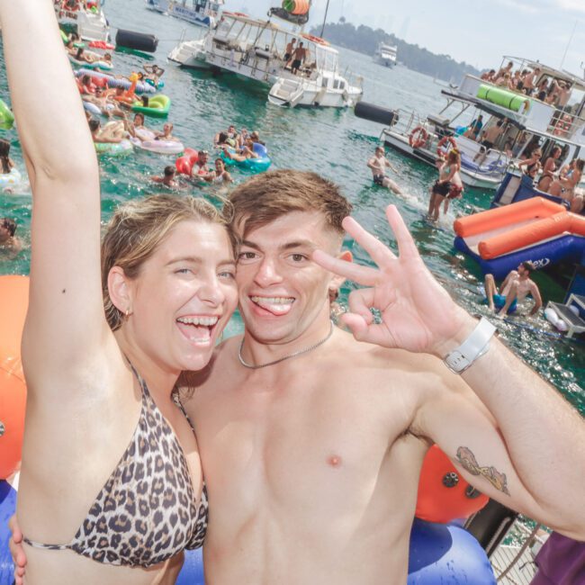 A smiling young woman in a leopard-print bikini and a shirtless man pose together, making peace signs, at a lively boat party with many people swimming and relaxing on floats in the water. Several boats are anchored nearby.
