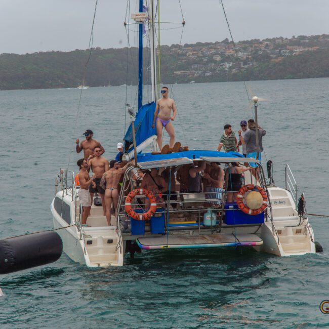 A group of people in swimwear are gathered on the deck of a catamaran in a bay, some posing and raising their arms, with forested hills and houses visible in the background under overcast skies.