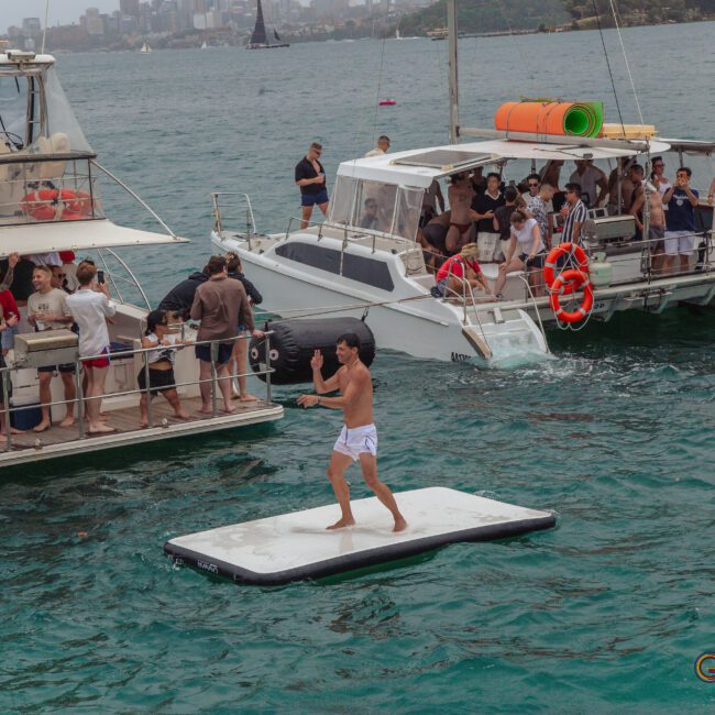 A man in swim trunks stands on a floating mat in the water between two boats filled with people socializing and watching him. The city skyline is visible in the background across the bay.