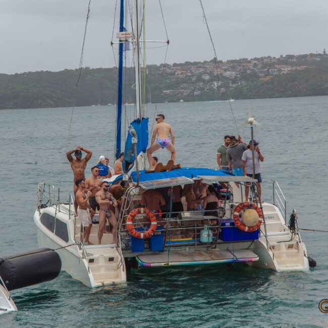 A group of people in swimsuits are gathered on a catamaran, some dancing and posing, with water and a tree-covered shoreline in the background under an overcast sky.