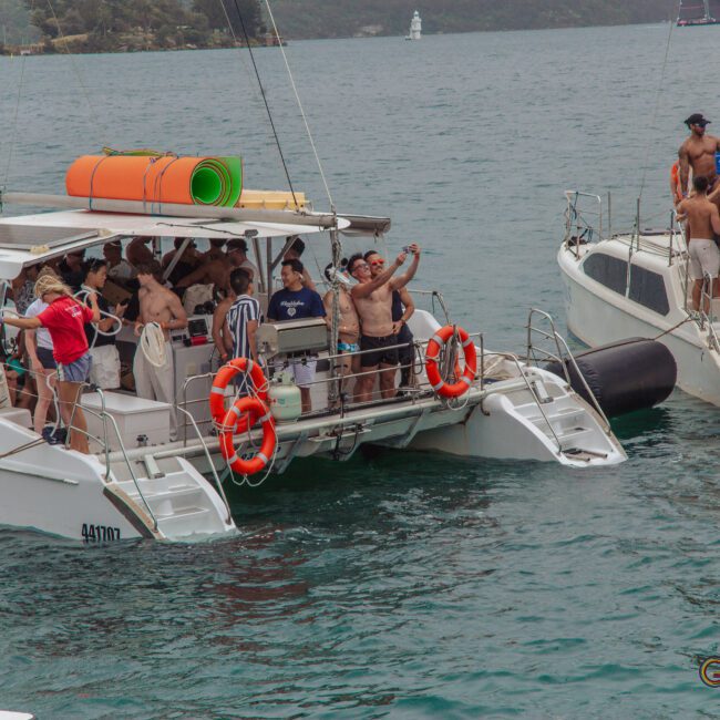 A group of people, many in swimwear, gather and take selfies on two docked catamarans in a tropical bay, with yoga mats and life rings visible. Lush green hills and other boats are seen in the background.