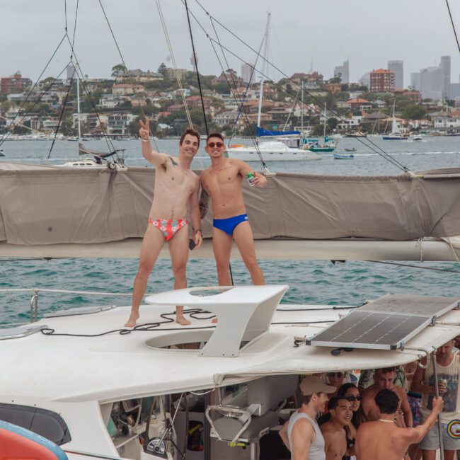 Two men in colorful swim briefs stand and pose on the roof of a boat, smiling and making peace signs, with other people gathered below deck. The boat is on the water with buildings and boats in the background.