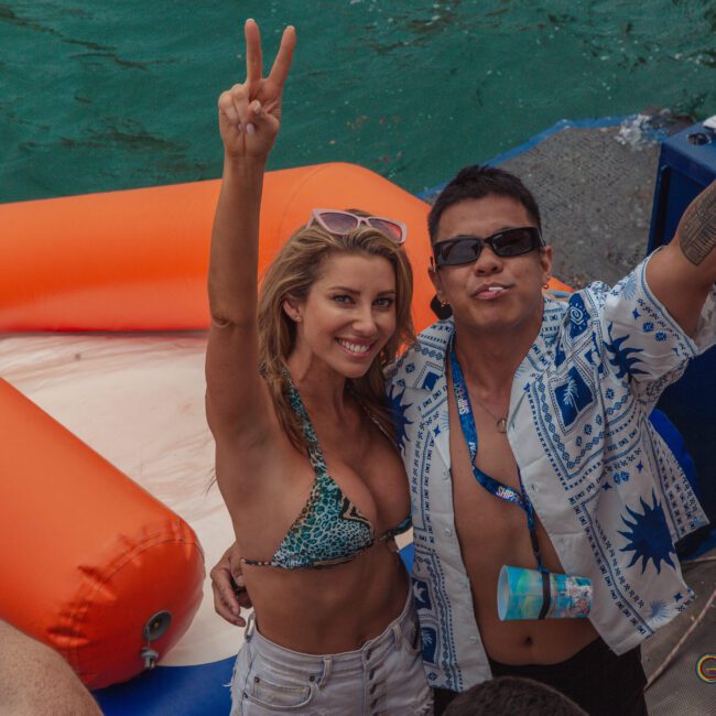 A smiling woman in a bikini and a man in sunglasses and a patterned shirt stand on a boat, both holding up peace signs. An orange inflatable float and green water are in the background.