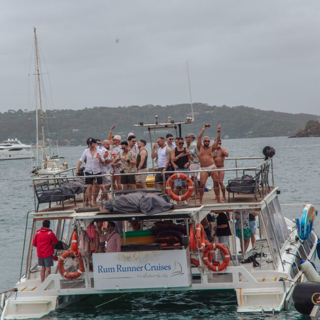 A group of people stand on the deck of a boat labeled "Rum Runner Cruises," cheering and posing for a photo on a cloudy day. The boat is on the water near a coastline with hills and other boats visible in the background.