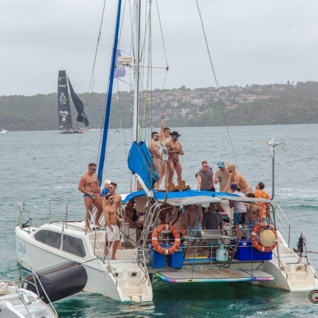 A group of people in swimsuits and hats are standing and sitting on a catamaran boat in a bay, celebrating. Other boats and a tree-covered shoreline are visible in the background on a cloudy day.